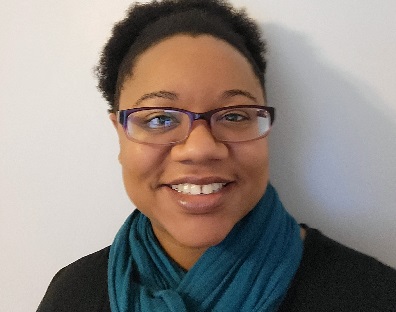 Picture of Ashley, a Black woman with hair up in a fro, wearing purple rimmed classes, smiling, and wearing a dark turquoise scarf and a black shirt.